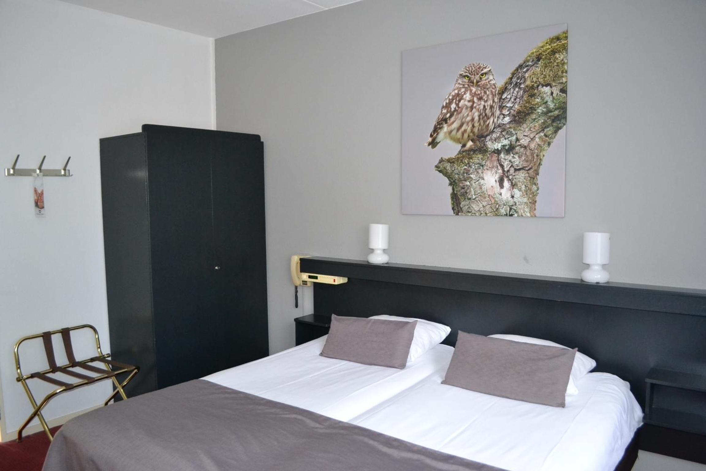 Et hotelværelse med en seng, en sort garderobe, et billede af en ugle på væggen og et lille bord med en lampe.