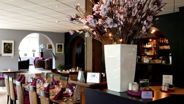 A restaurant with a long dining table, chairs, and a large vase of flowers. - Veluwe Hotel Stakenberg (Elspeet)