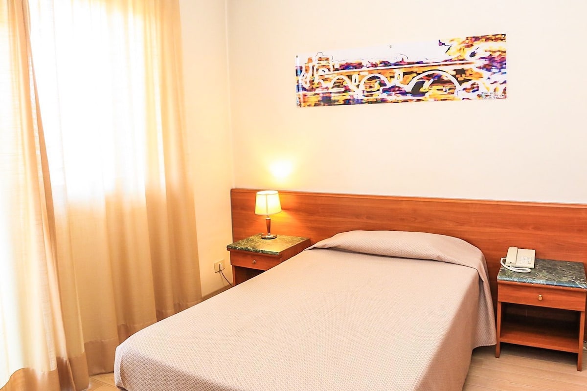A hotel room with a bed, a nightstand with a lamp, a telephone, and a painting on the wall.