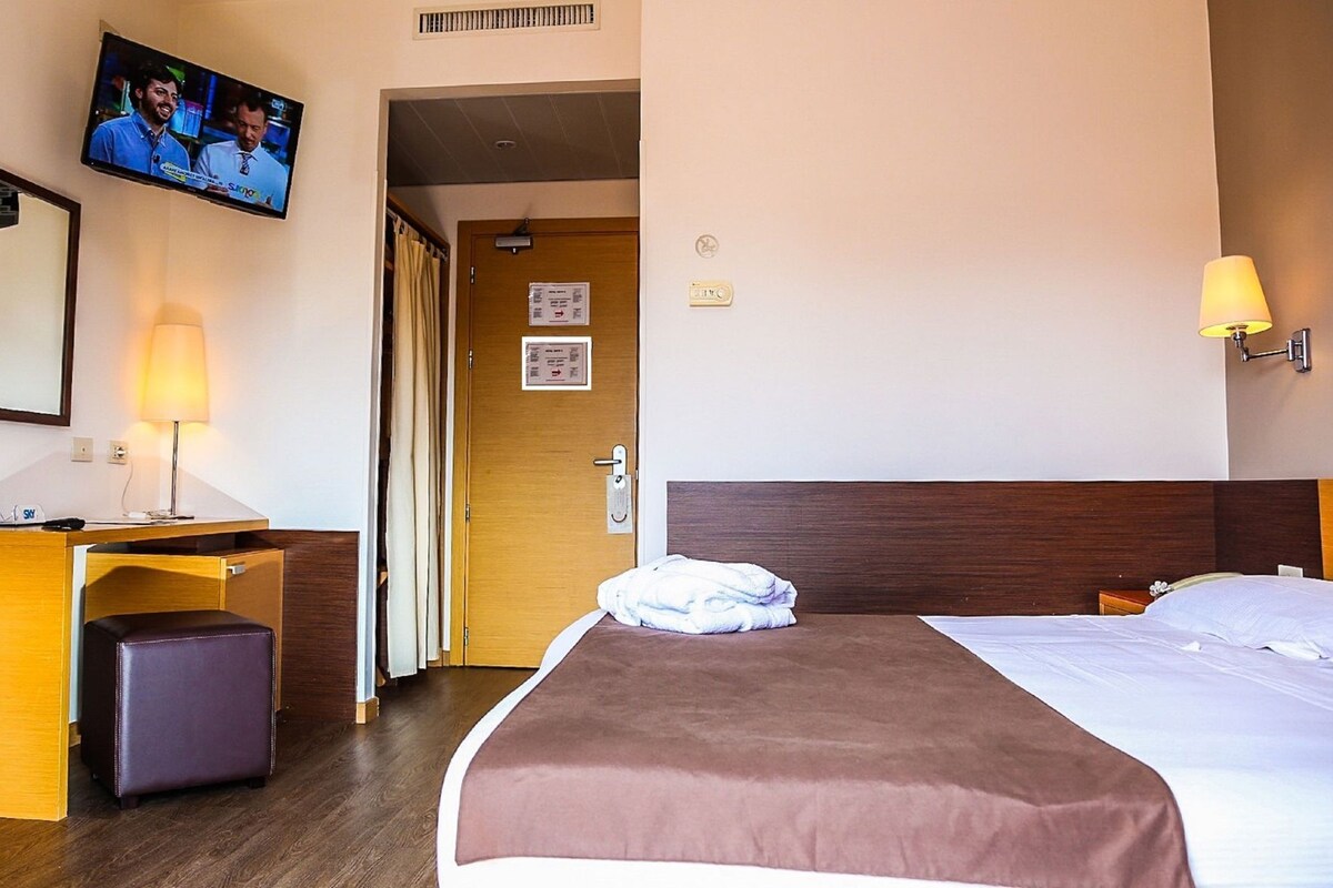 A hotel room with a bed, a desk with a lamp, a television on the wall, and a door.
