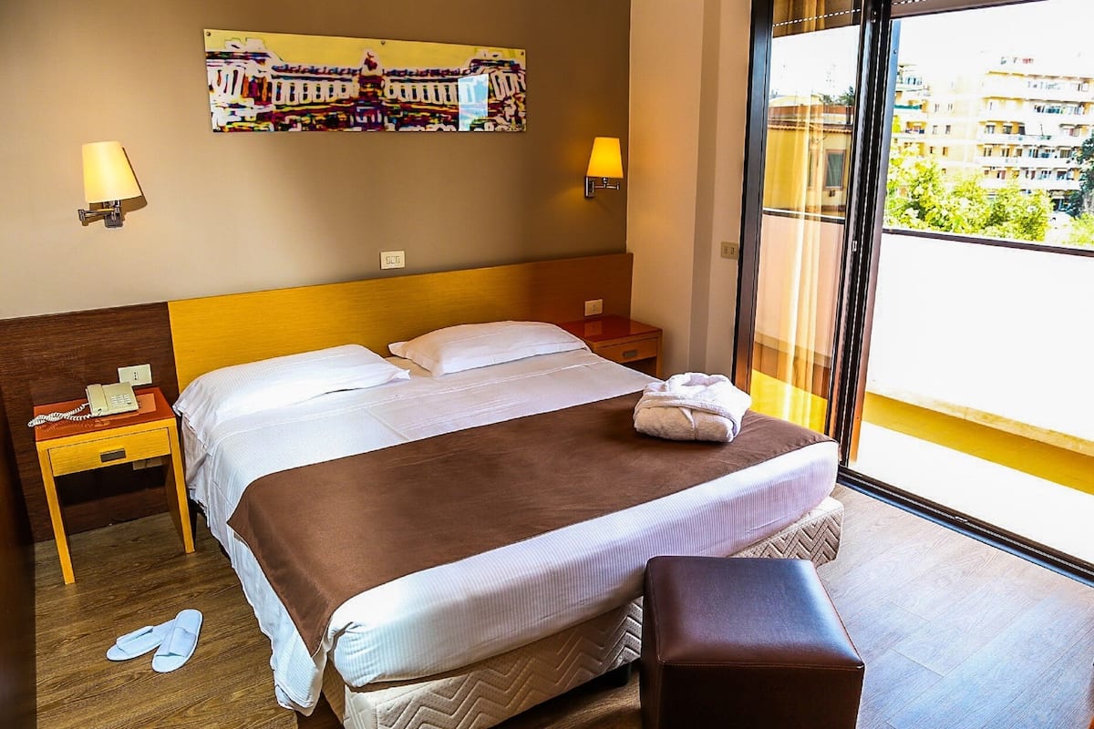 A hotel room with a large bed, a bedside table with a phone, a nightstand, a window with a view of buildings, and a wall-mounted artwork.