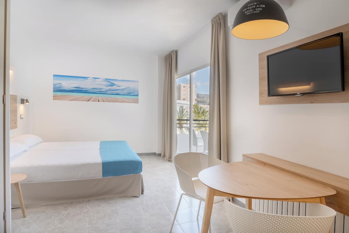 Ein Hotelzimmer mit einem Bett, einem Tisch, einem Sessel, einem Fernseher und einem Strandbild.