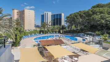 Un área de piscina en la azotea con sillones de descanso y sombrillas, rodeada de palmeras y edificios.