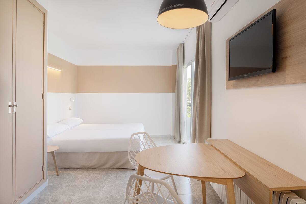 Ein modernes Hotelzimmer mit Bett, Schreibtisch, Stuhl, Wandfernseher und einem Vorhangfenster.