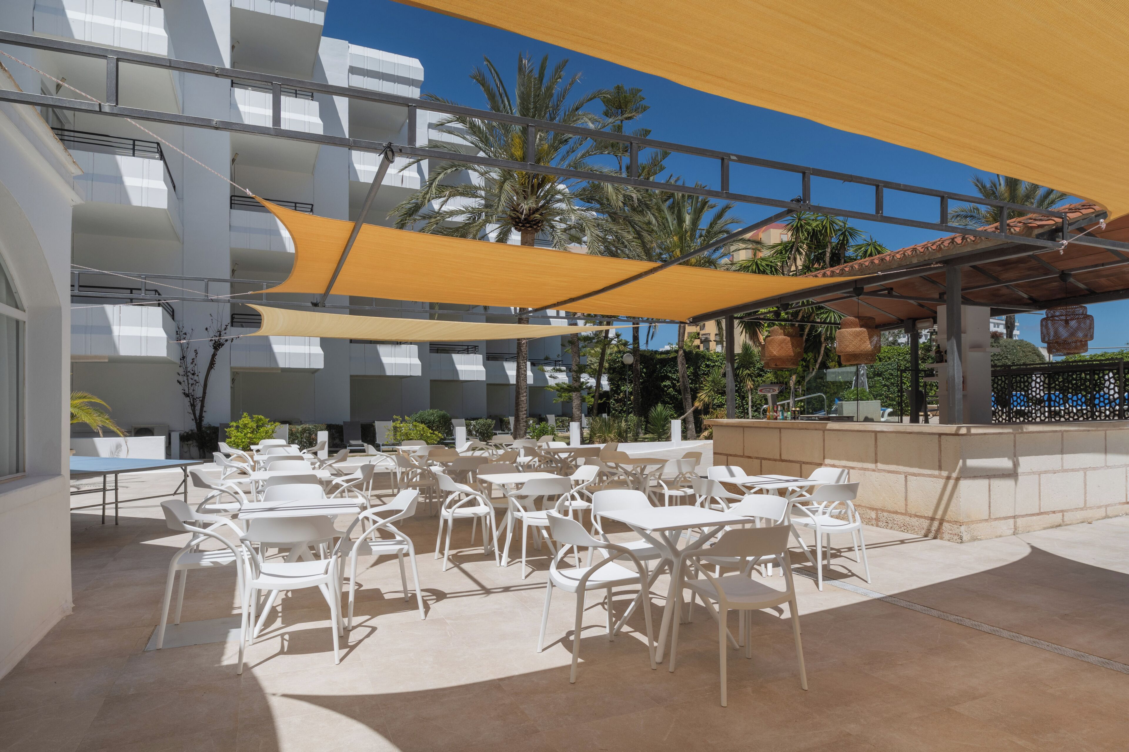 Un área de estar al aire libre con sillas y mesas blancas, protegida por un amplio toldo amarillo, rodeada de palmeras y un edificio moderno.
