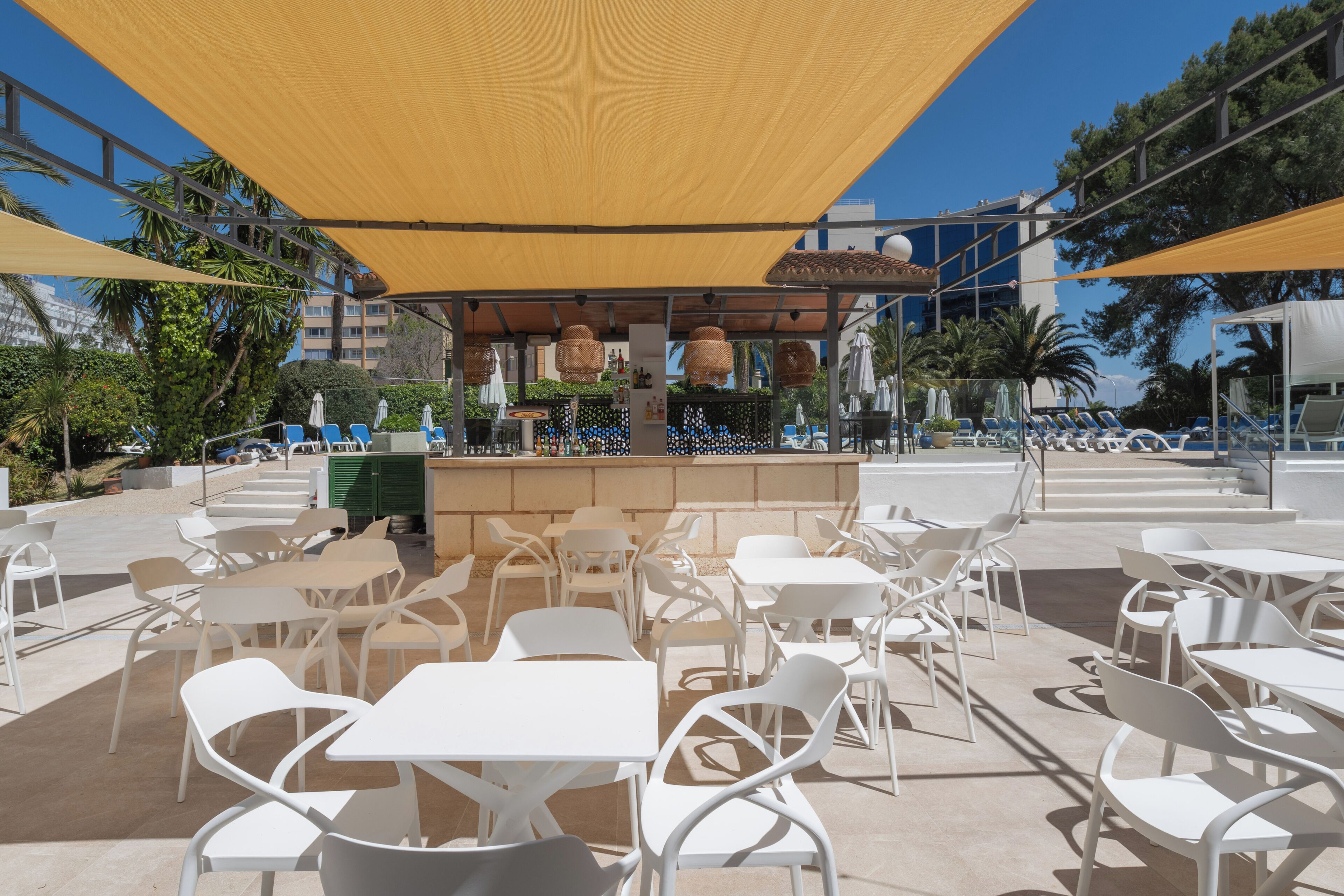 An outdoor seating area with white tables and chairs, a bar with hanging lights, and umbrellas providing shade.