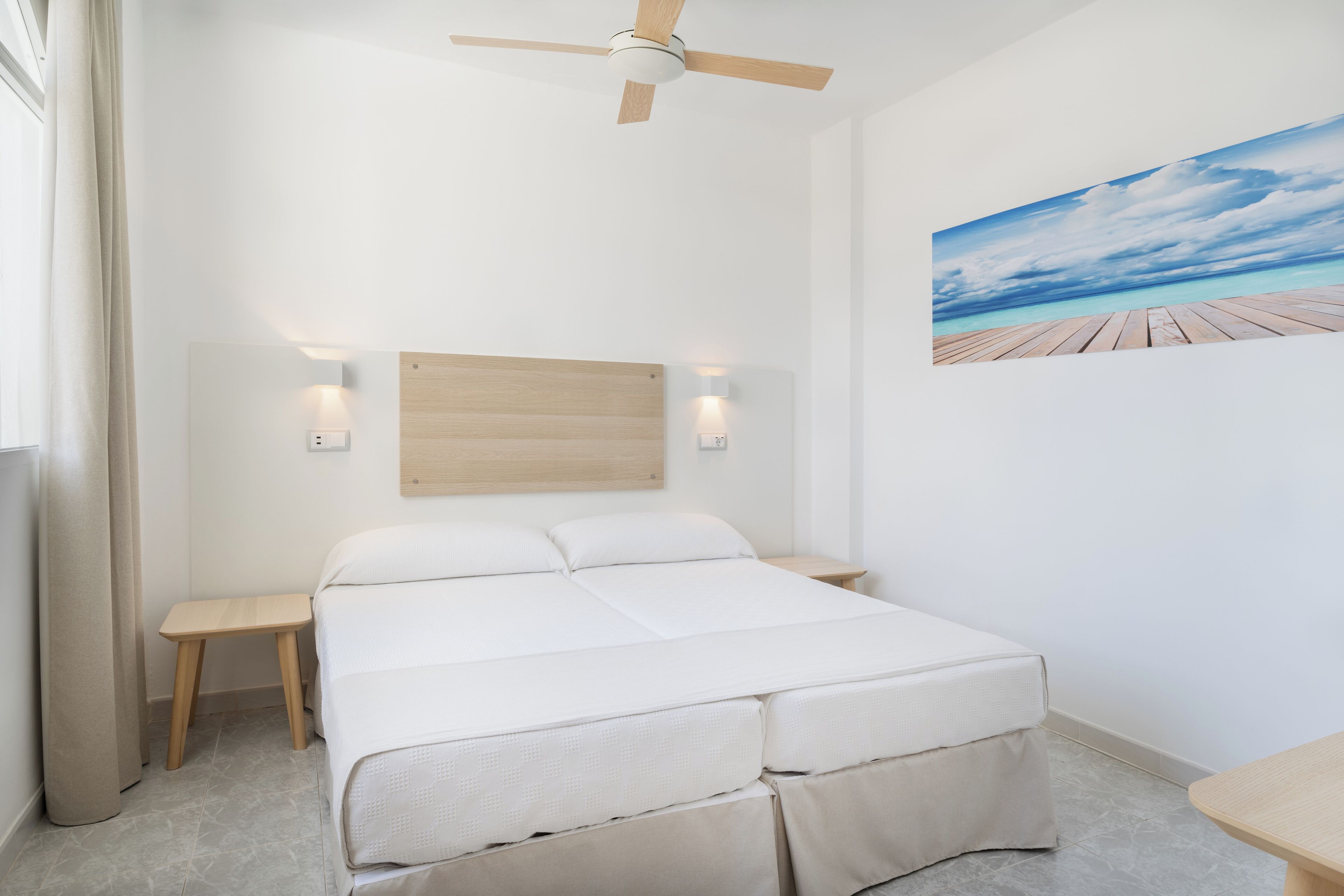 A bedroom with a bed, a ceiling fan, a nightstand, a wall-mounted light, and a painting of a beach scene.