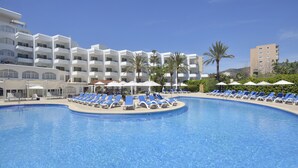 A large swimming pool with blue lounge chairs and umbrellas, surrounded by a white building with multiple balconies and palm trees.
