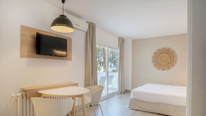 A modern hotel room with a flat-screen TV mounted on a wooden panel, a round dining table with white chairs, a white sofa, and a large window with curtains.