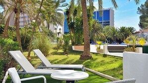 A poolside area with lounge chairs and a small table, surrounded by palm trees and greenery, with a building in the background.