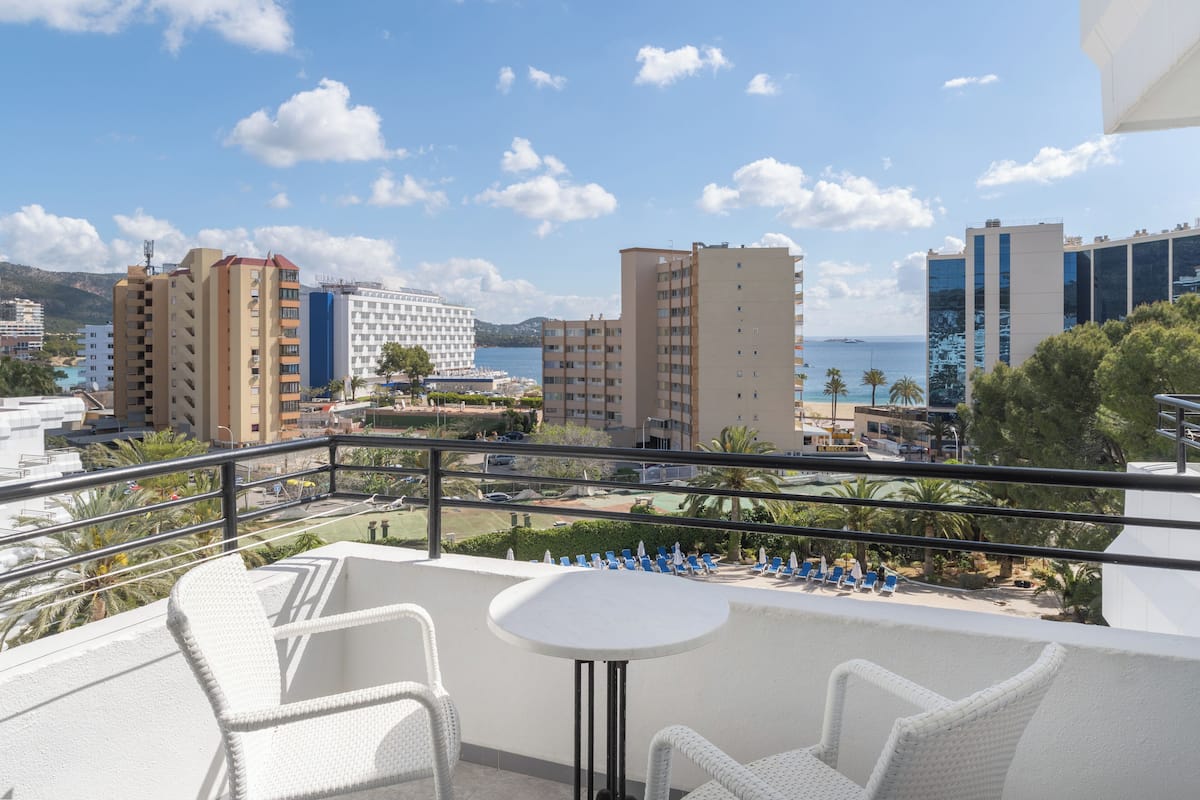 Ein Balkon mit weißen Korbmöbeln, einem kleinen runden Tisch und Blick auf das Meer und die Gebäude.
