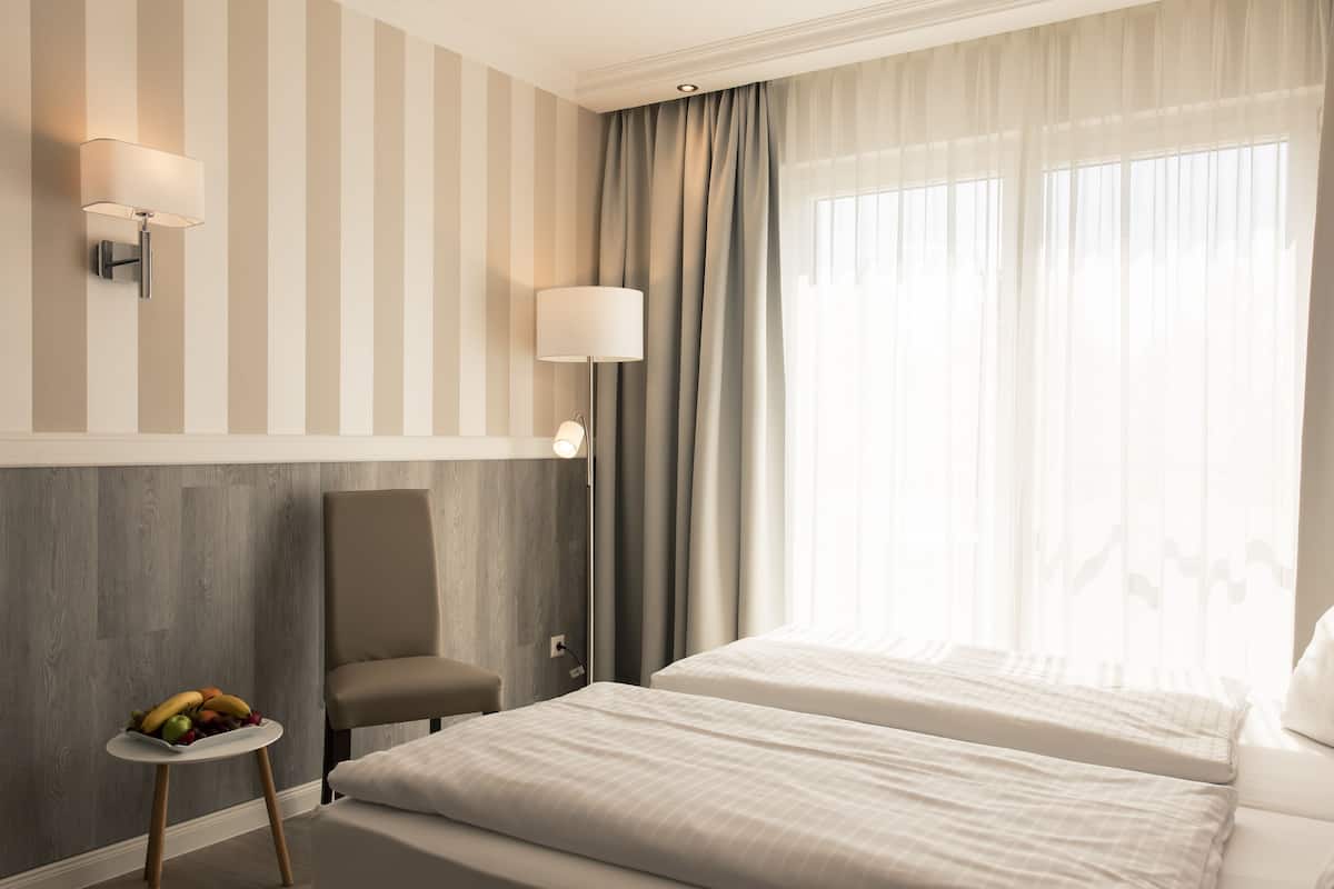 Ein Hotelzimmer mit zwei Betten, einem kleinen Tisch mit Obst, einem Sessel, einer Lampe und einem Fenster mit Vorhängen.