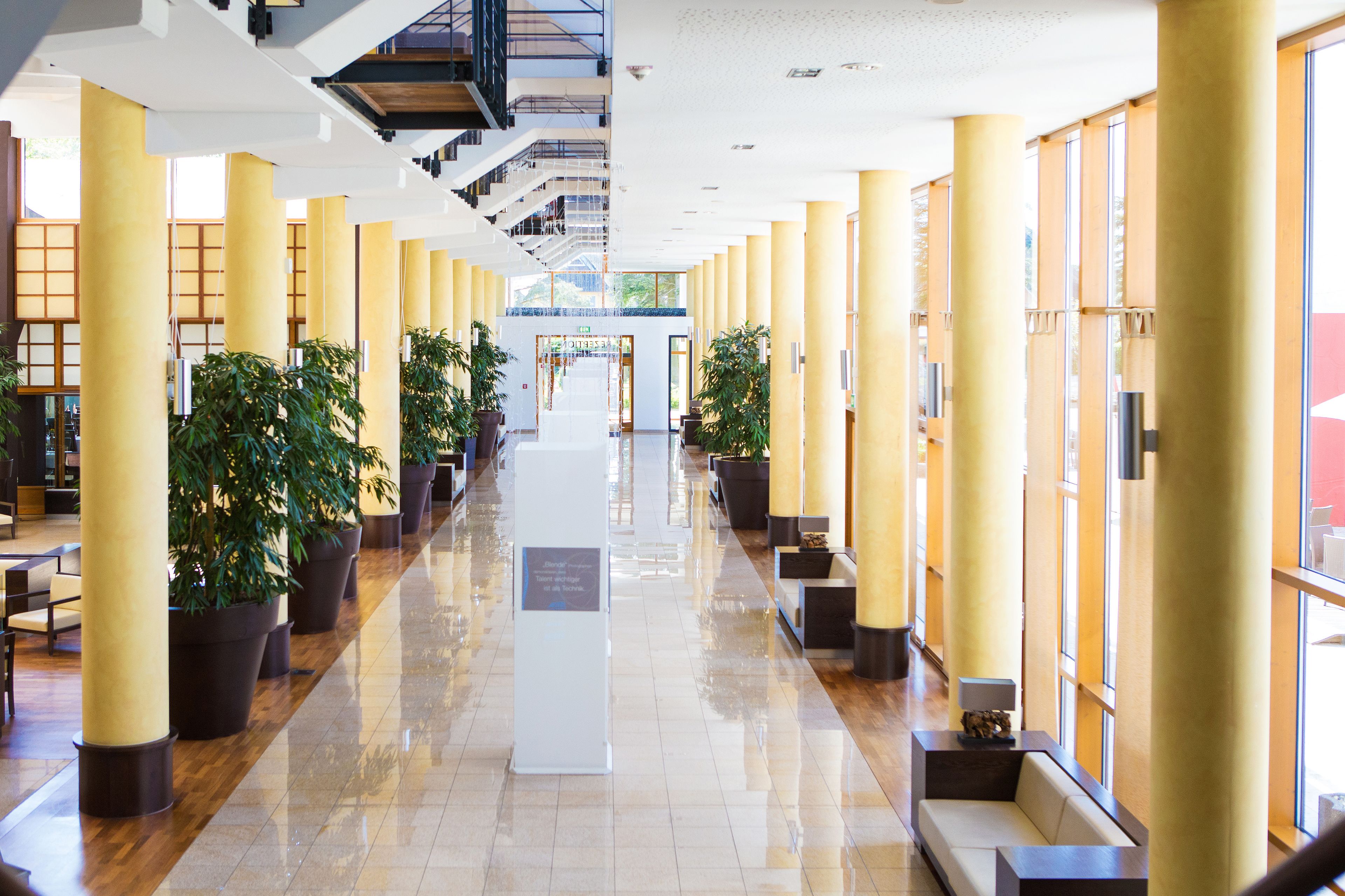 A spacious hotel lobby with a central walkway flanked by large potted plants and yellow pillars.
