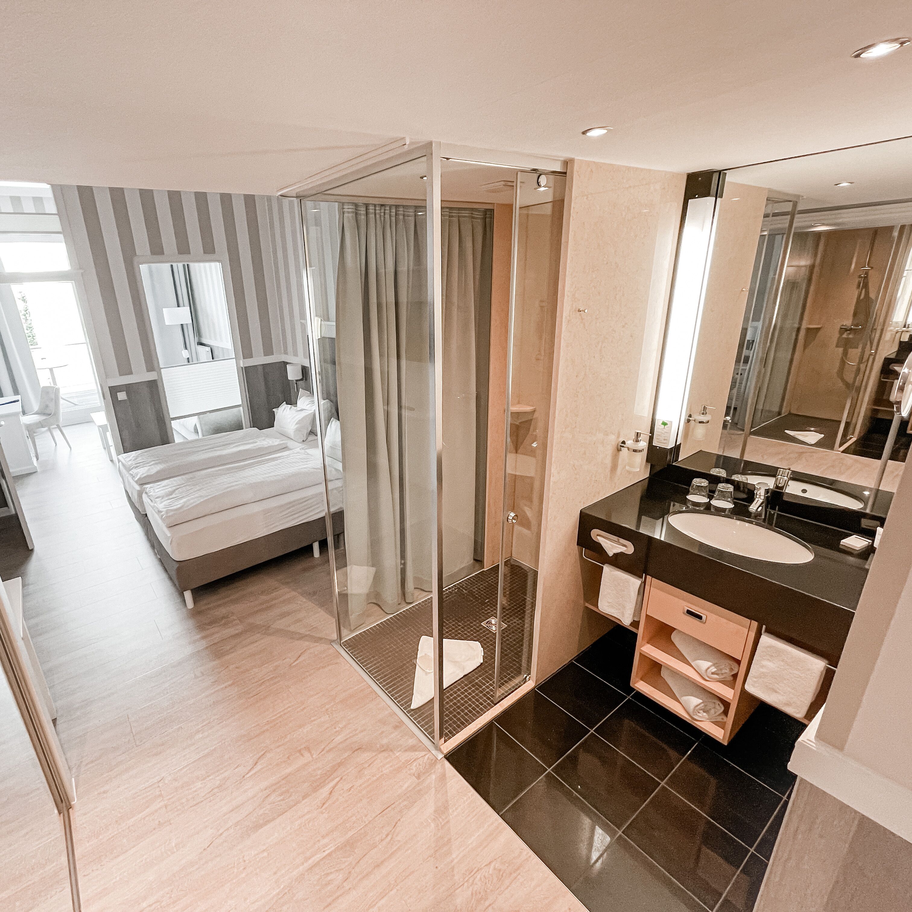 Una habitación de hotel moderna con cama, ducha con paredes de vidrio y baño con encimera negra y lavamanos blanco.
