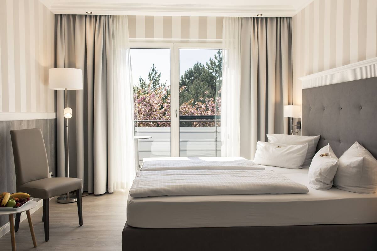 Ein Hotelzimmer mit einem großen Bett, einem Sessel, einem Tisch mit Obst und Blick auf Bäume durch das Fenster.