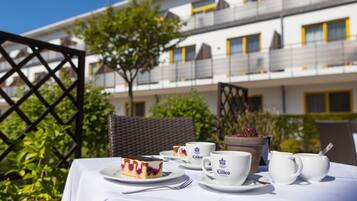 Ein Tisch ist gedeckt mit einem Kuchenstück, zwei Kaffeetassen und einem Teekessel, im Hintergrund ein Balkon und Grünpflanzen.