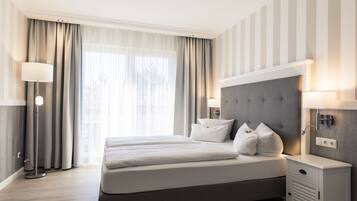 A modern hotel room with a large bed, two bedside lamps, a nightstand, and a view of the outdoors through sheer curtains.