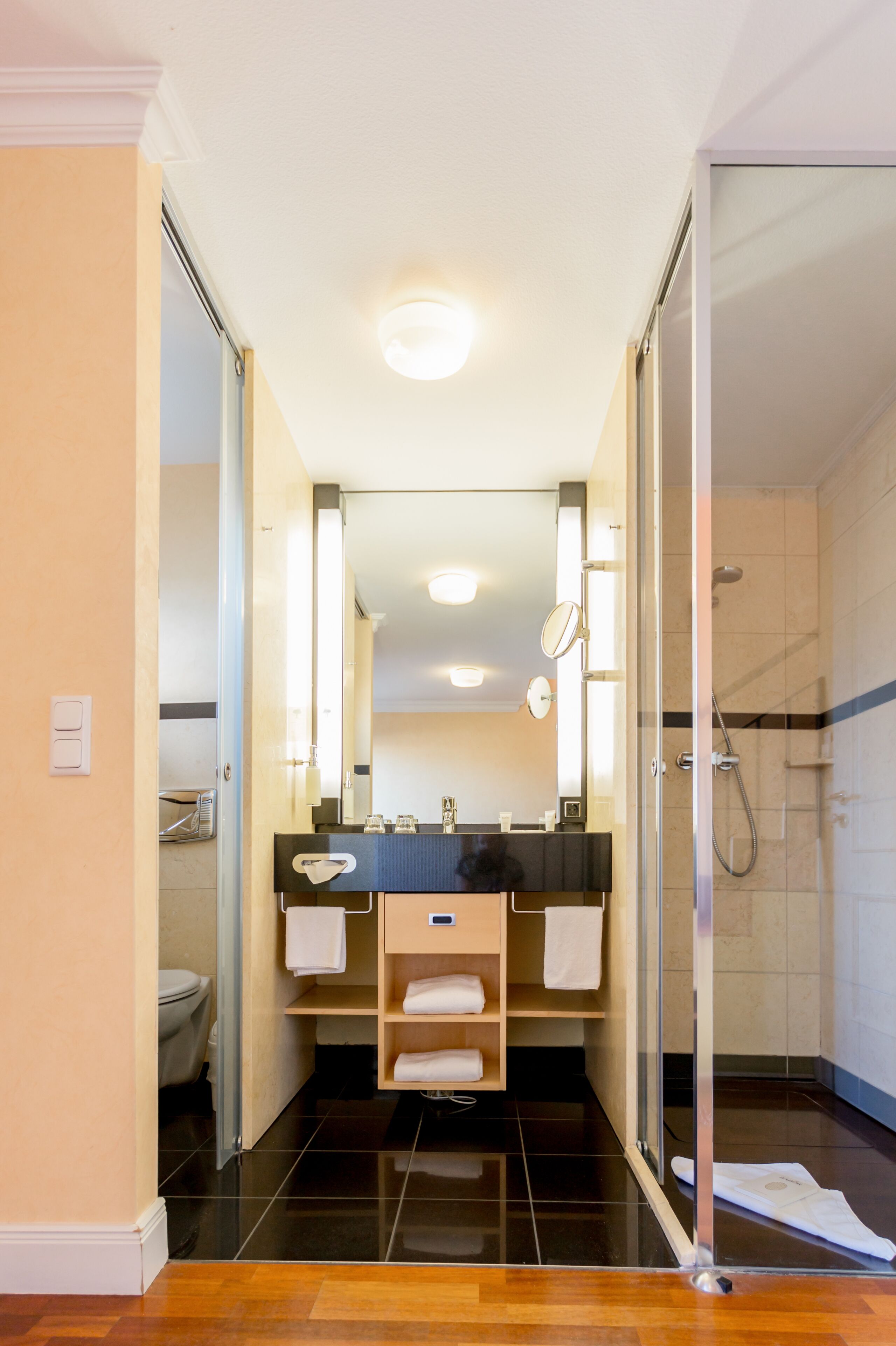 Ein modernes Badezimmer mit Duschkabine aus Glas, einem Doppelwaschtisch und einer Toilette, die durch eine offene Tür sichtbar ist.