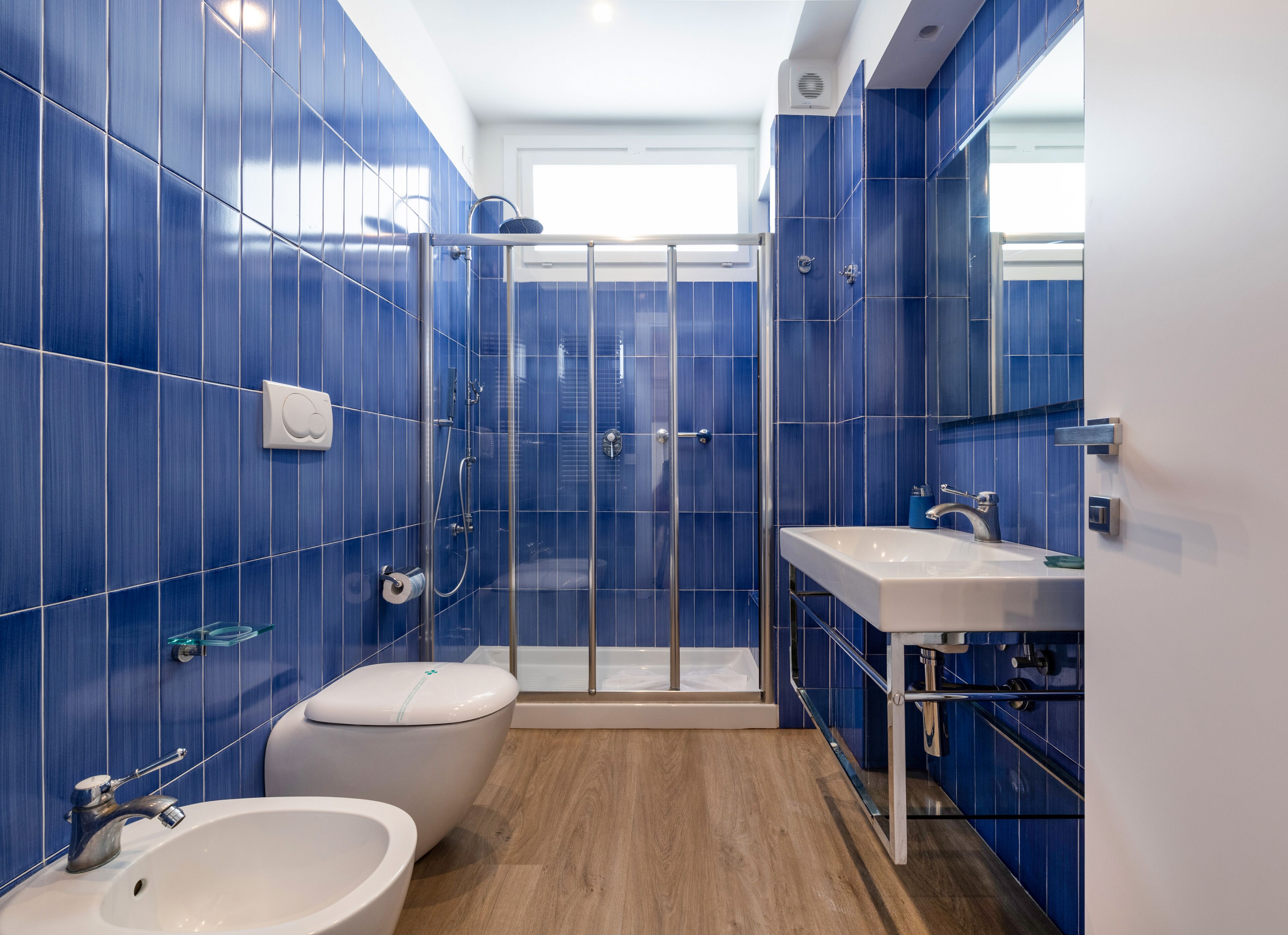 Un baño moderno con azulejos azules, una cabina de ducha de cristal, un inodoro, un lavabo y suelo de madera.