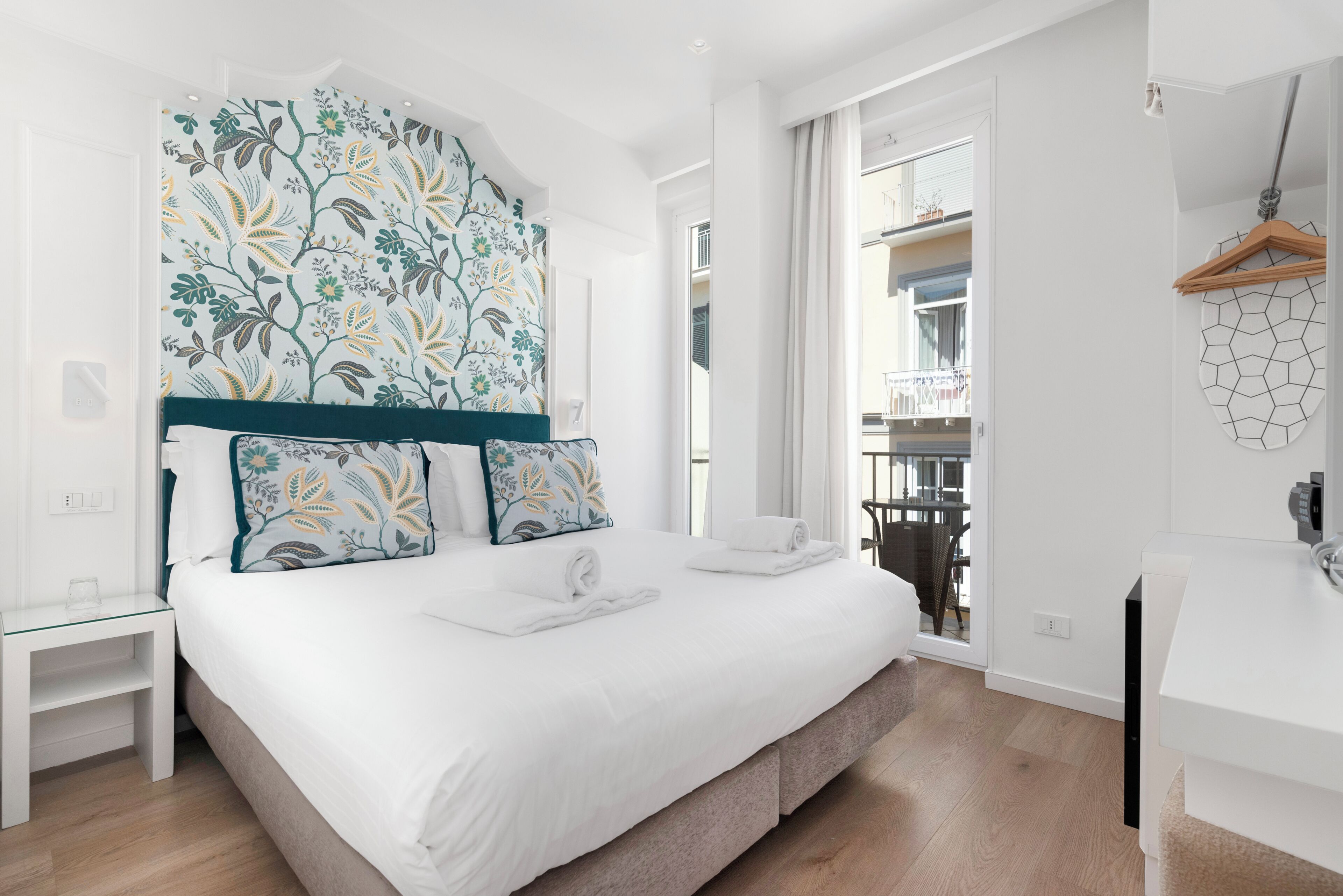 A modern hotel room with a large bed, a view to the outside, a small desk, and a wardrobe.