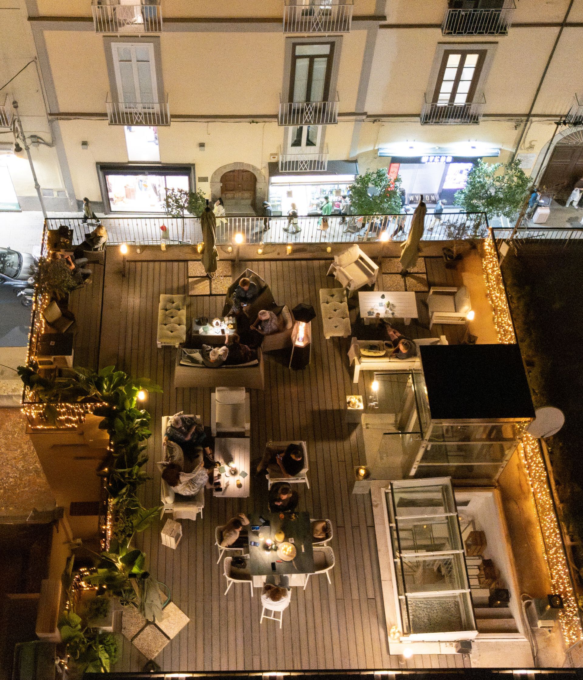 An outdoor seating area with multiple tables and chairs, surrounded by plants and string lights.