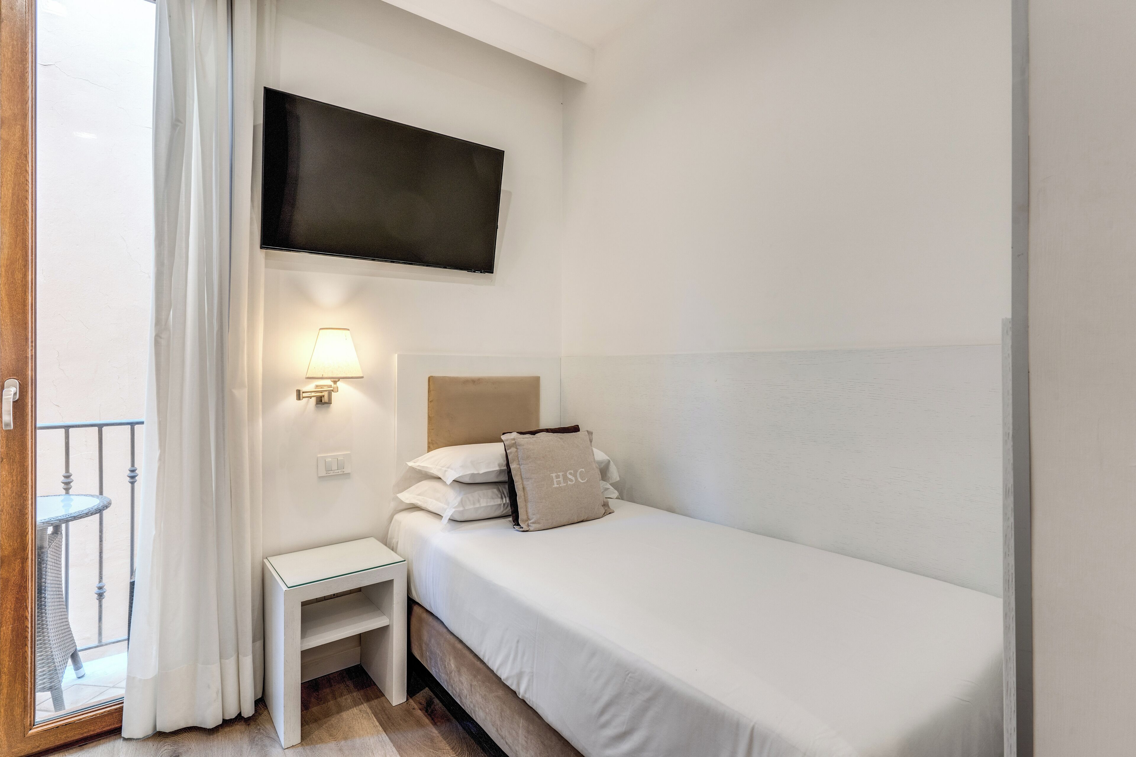A small, single-bed room with a TV mounted on the wall, a bedside table with a lamp, and a view of the outside balcony.