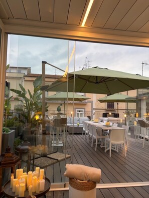 Een terras buiten met tafels en stoelen, een glazen scheiding en een uitzicht op gebouwen op de achtergrond.