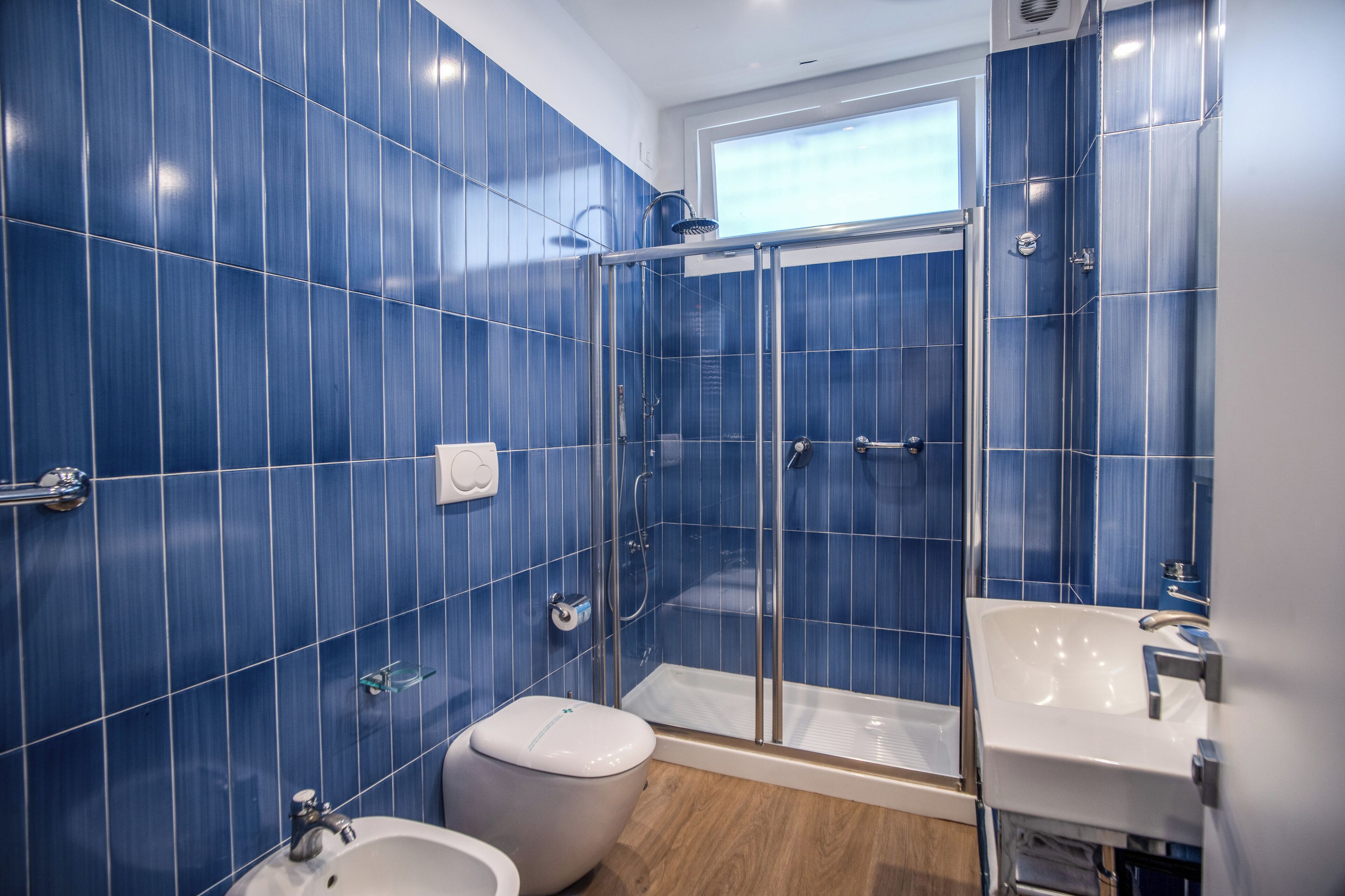 Un baño con azulejos azules, mampara de vidrio, lavamanos blanco y un inodoro.