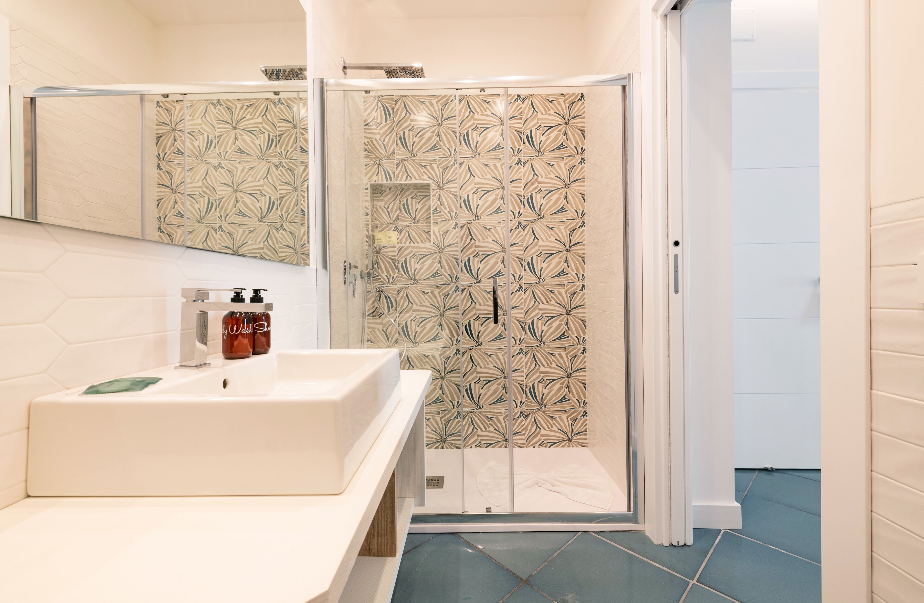 Un baño con lavamanos blanco, un espejo y una ducha con pared de azulejos estampados.