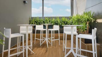 Un área de estar moderna al aire libre con sillas y mesas blancas, rodeada de plantas en macetas y amplias ventanas que ofrecen vistas del cielo.