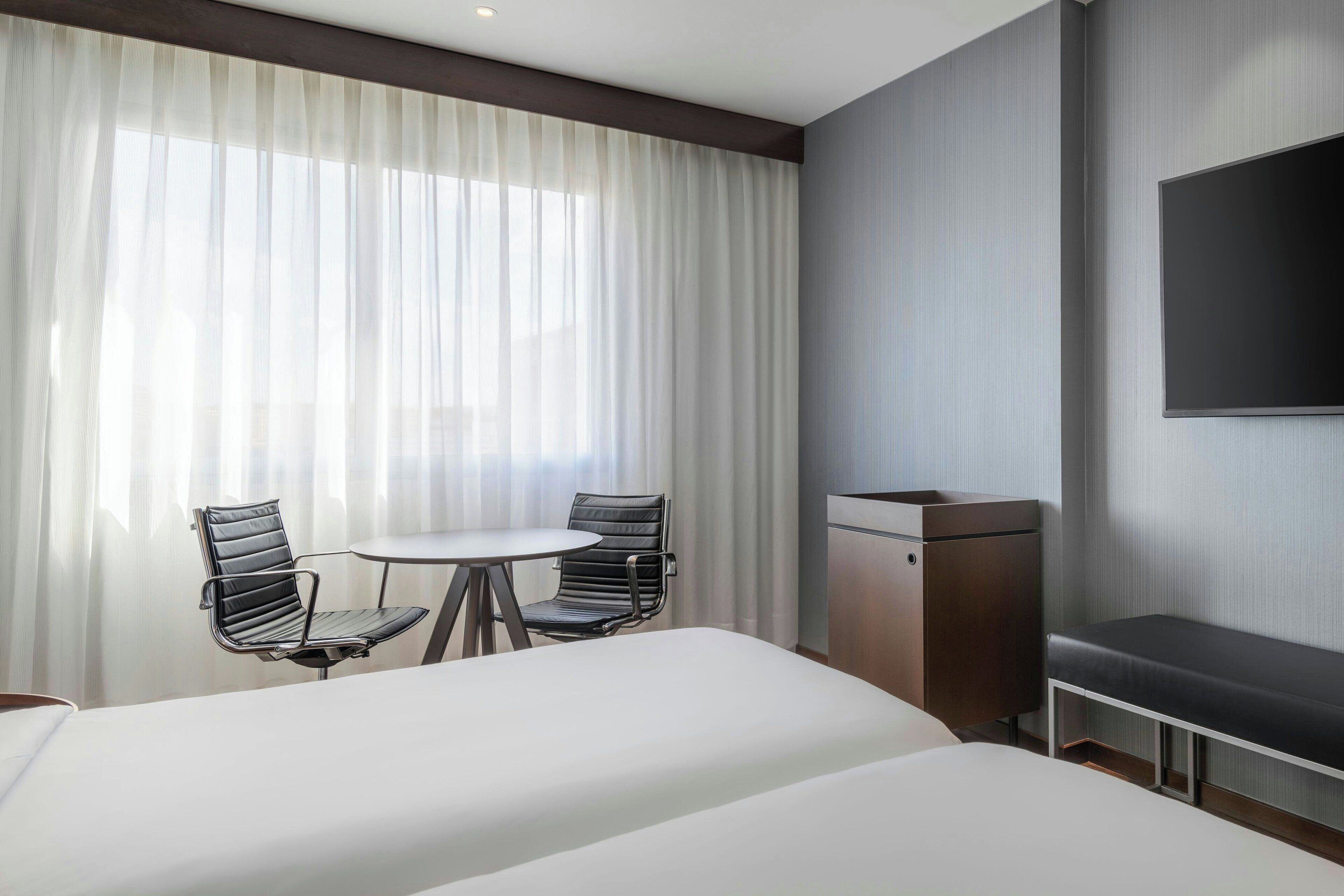Une chambre d’hôtel avec deux lits, une petite table avec des chaises, un téléviseur fixé au mur et une grande fenêtre avec des rideaux légers.