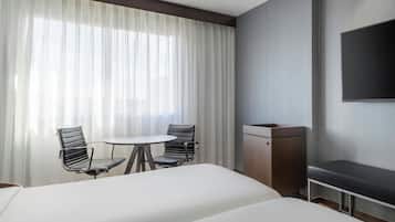 Una habitación de hotel con dos camas, una mesita con sillas, un televisor en la pared y un gran ventanal con cortinas traslúcidas.