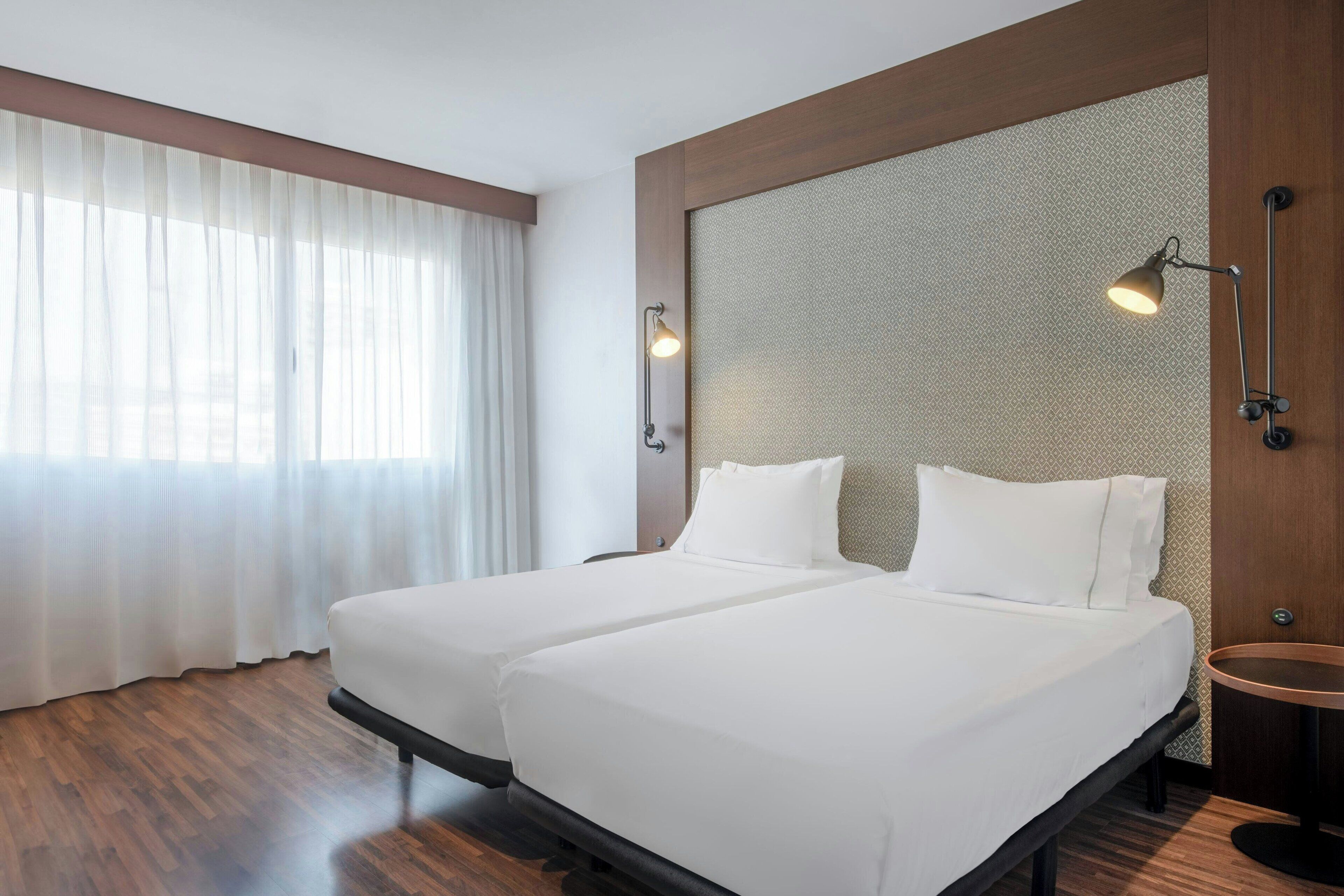 A hotel room with two beds, a large window with sheer curtains, a round table, and a textured wall panel.