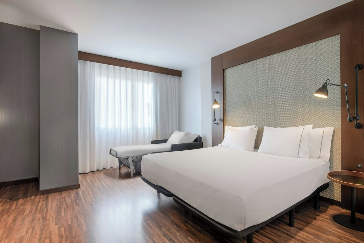 Una habitación de hotel moderna con una cama grande, una mesita de noche, una silla y un ventanal con cortinas.