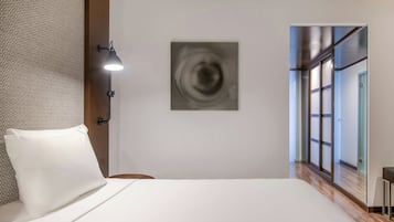 A hotel room with a bed, a wall-mounted lamp, a modern abstract painting, and a view of a corridor with glass doors.
