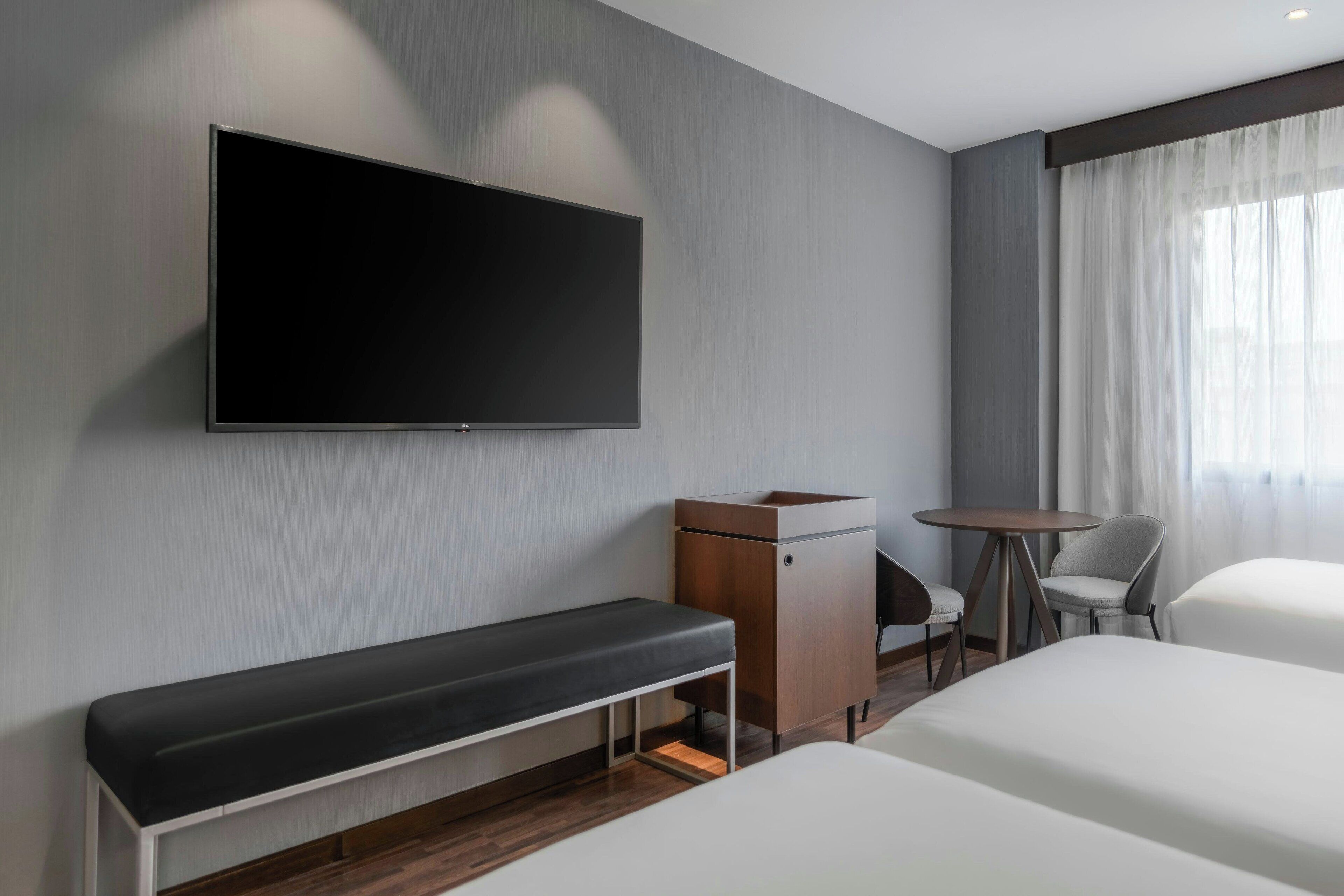 A modern hotel room with a large flat-screen TV mounted on the wall, a bench, a small desk, two chairs, and a bed.