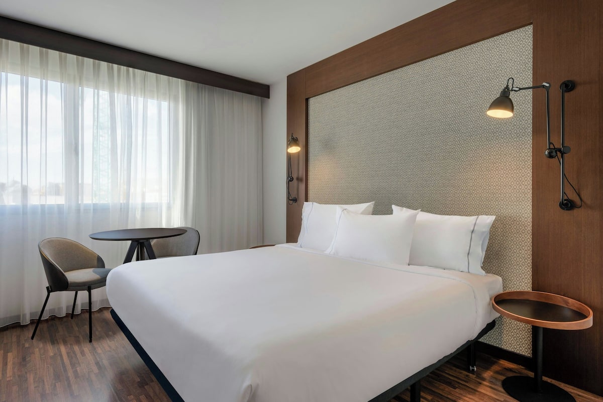 Una habitación de hotel con una cama grande, una mesita redonda pequeña, una silla y una lámpara fijada a la pared.