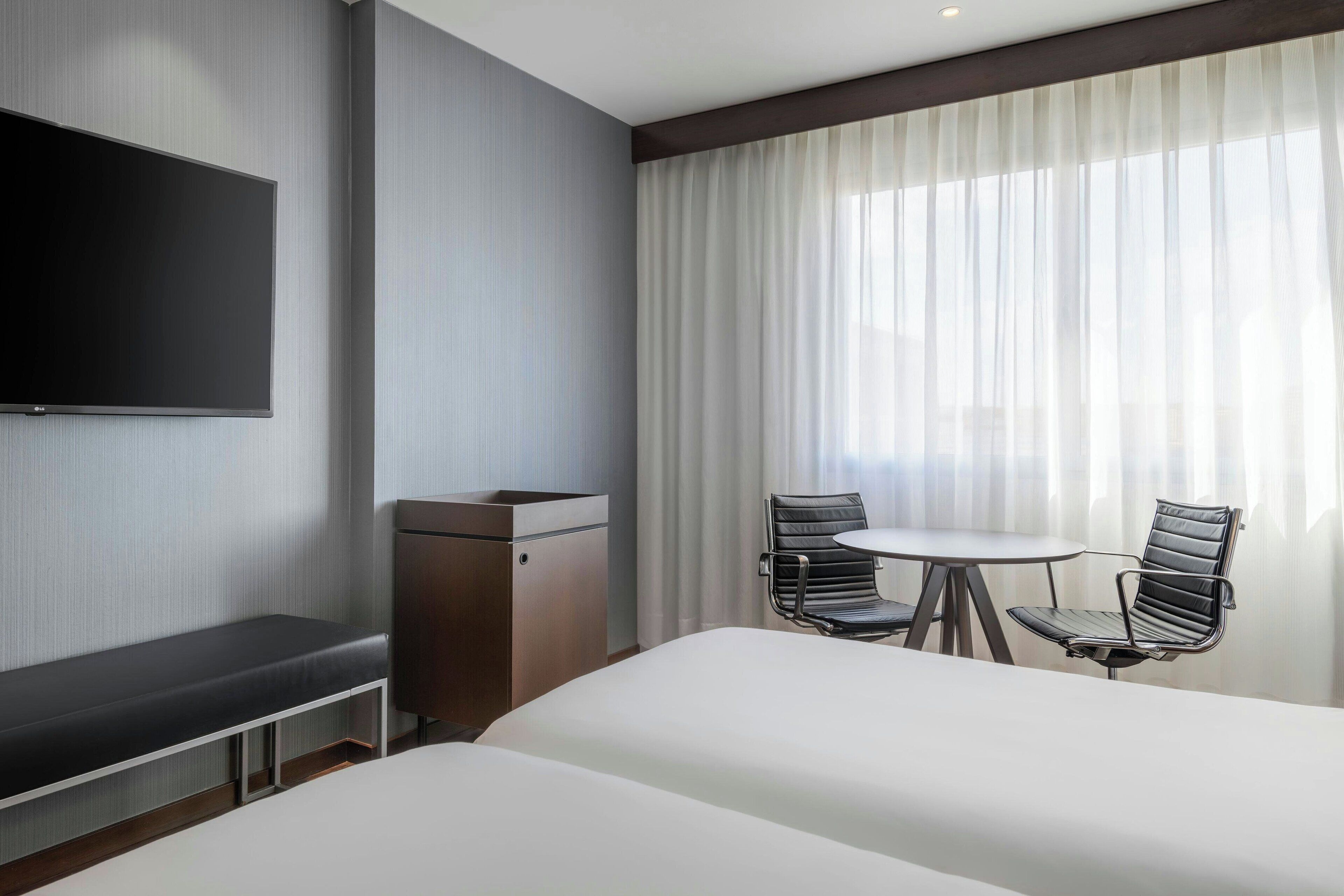 Una habitación de hotel con cama, escritorio, silla, televisor y ventana con cortinas.