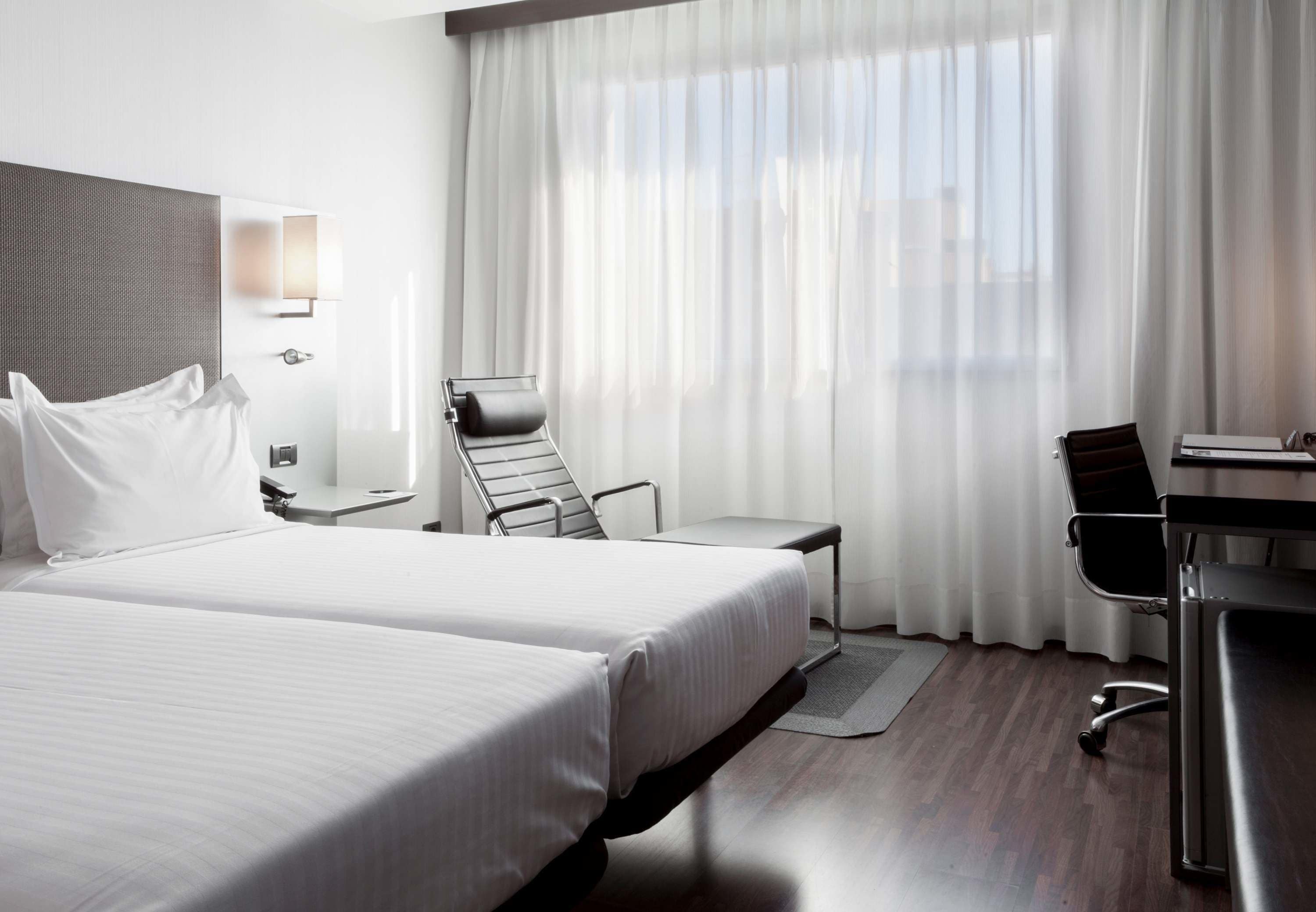 A hotel room with two beds, a desk, a chair, and a large window with sheer curtains.