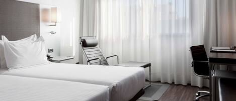 A hotel room with two beds, a desk, a chair, and a large window with sheer curtains.