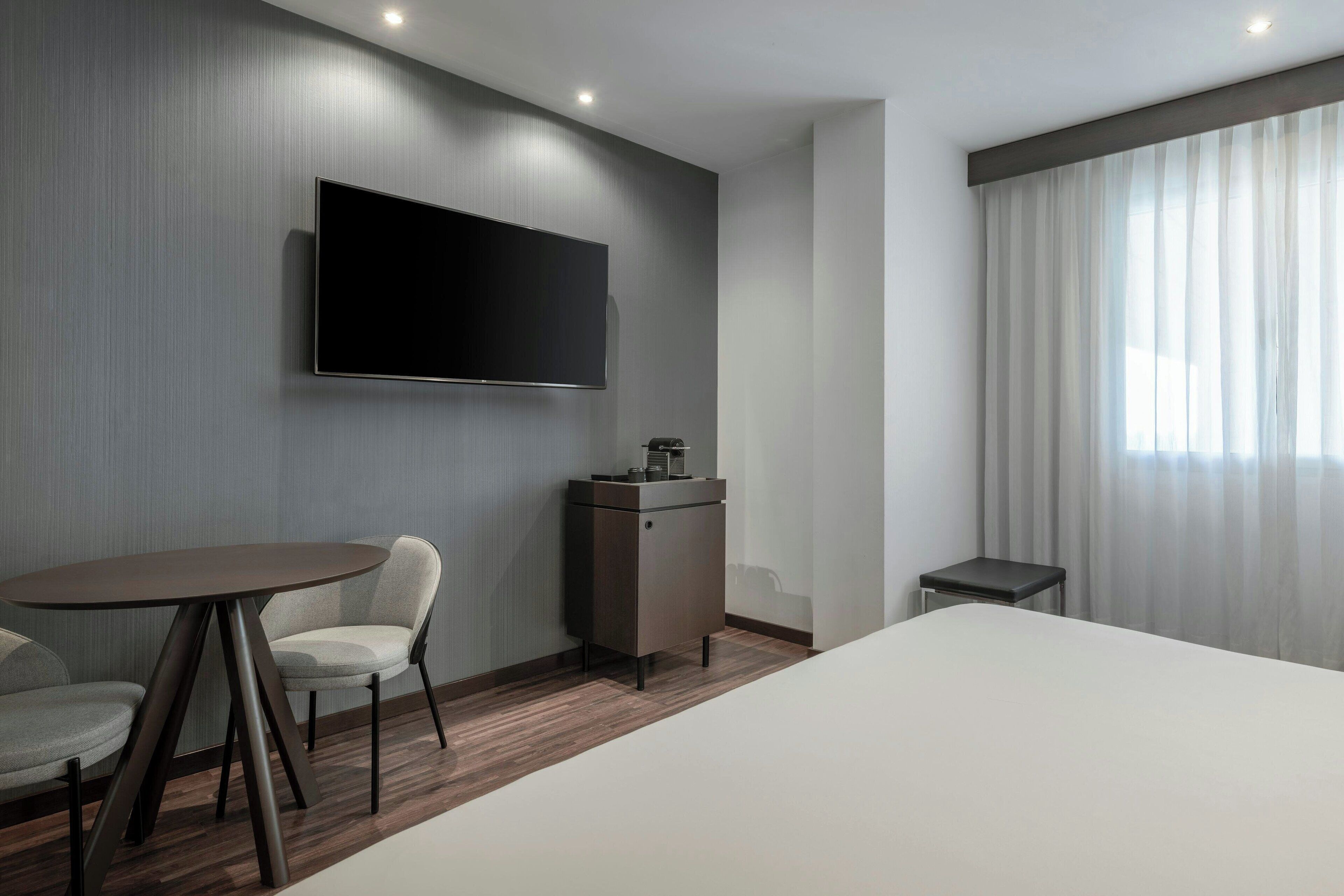 A modern hotel room with a flat-screen TV mounted on the wall, a small cabinet, a round table with two chairs, a bed, and a window with sheer curtains.
