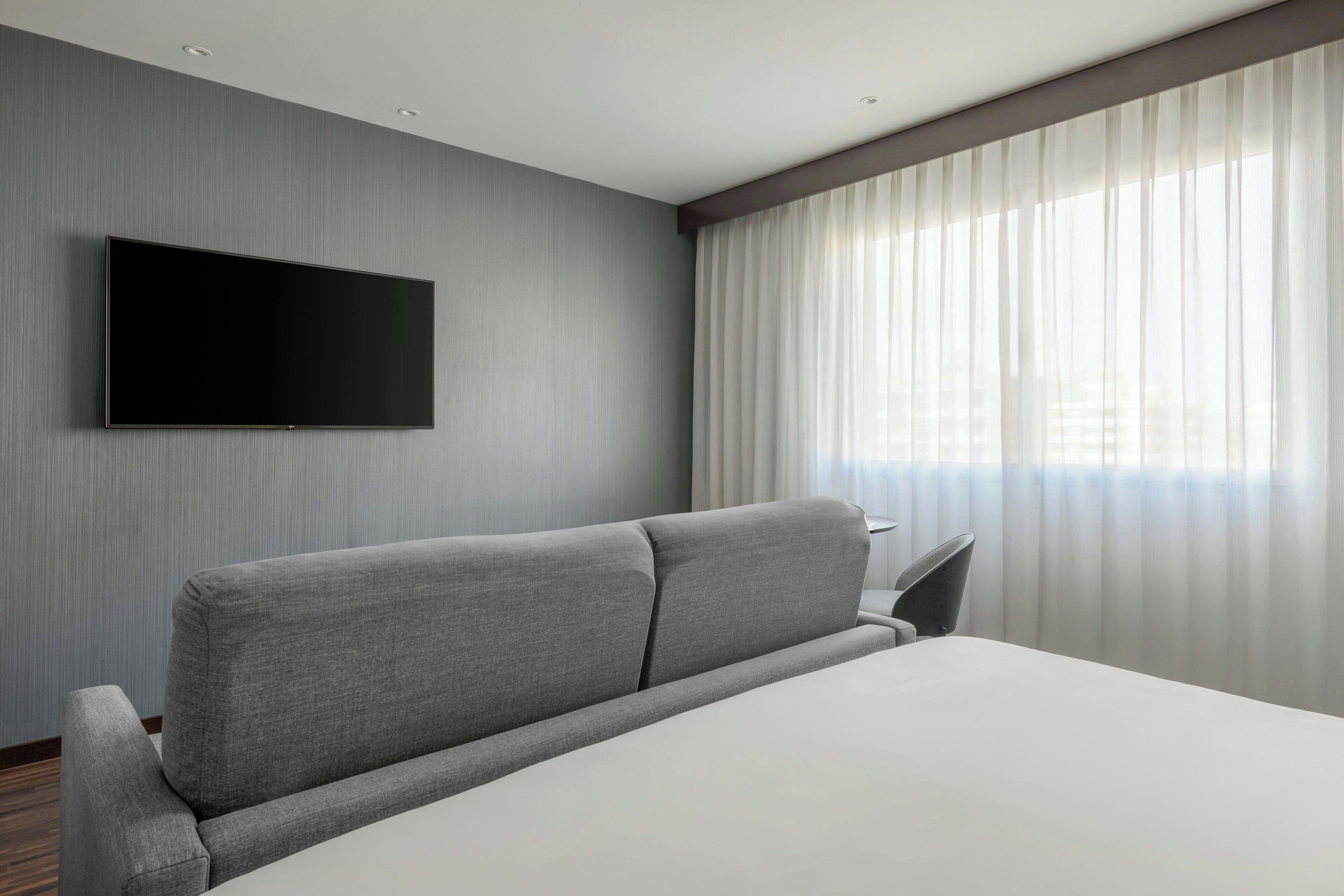A modern hotel room with a large bed, a flat-screen TV mounted on the wall, and a large window with sheer curtains.