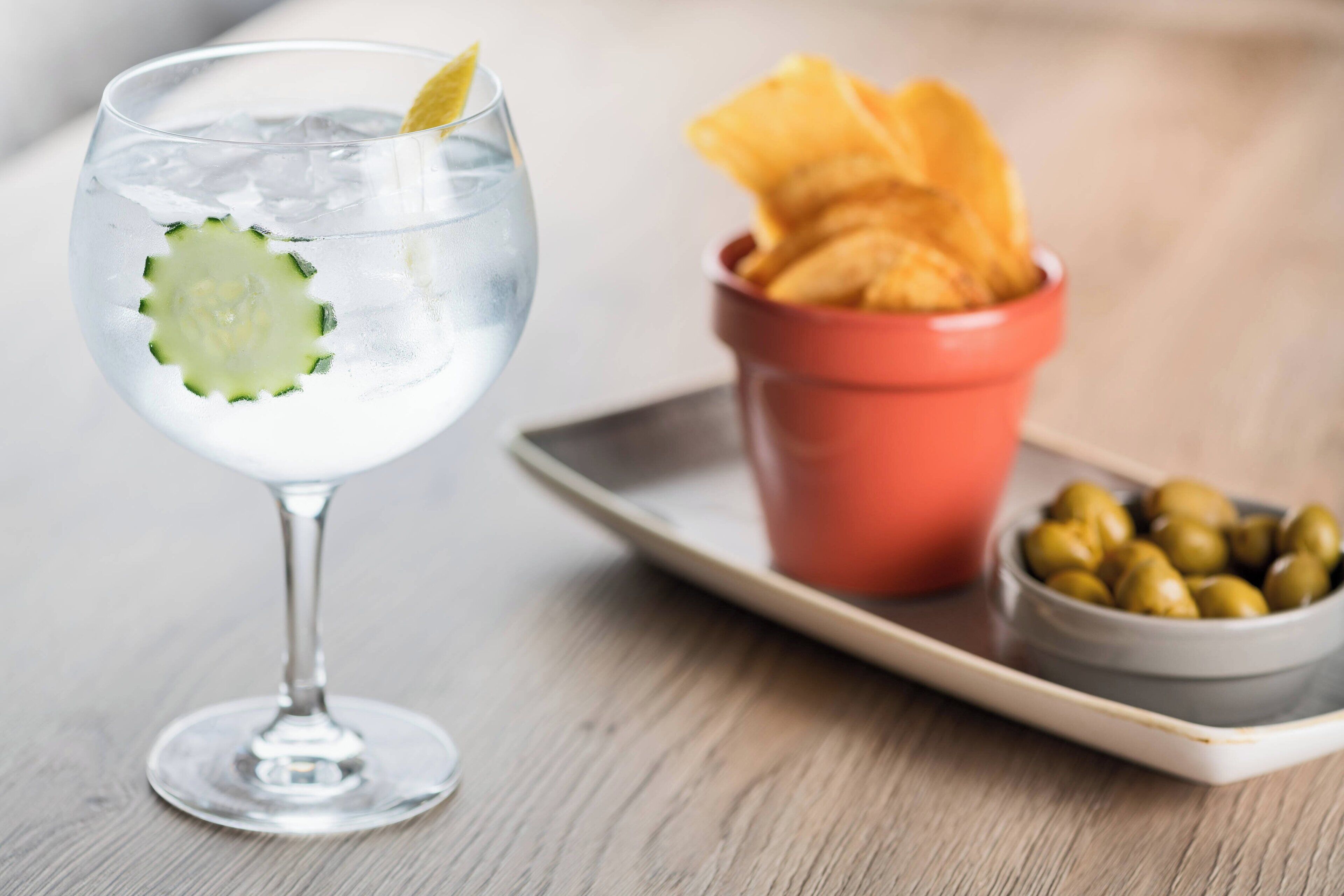 Een glas helder drankje met een plak komkommer, een klein schaaltje olijven en een bakje chips op een dienblad.