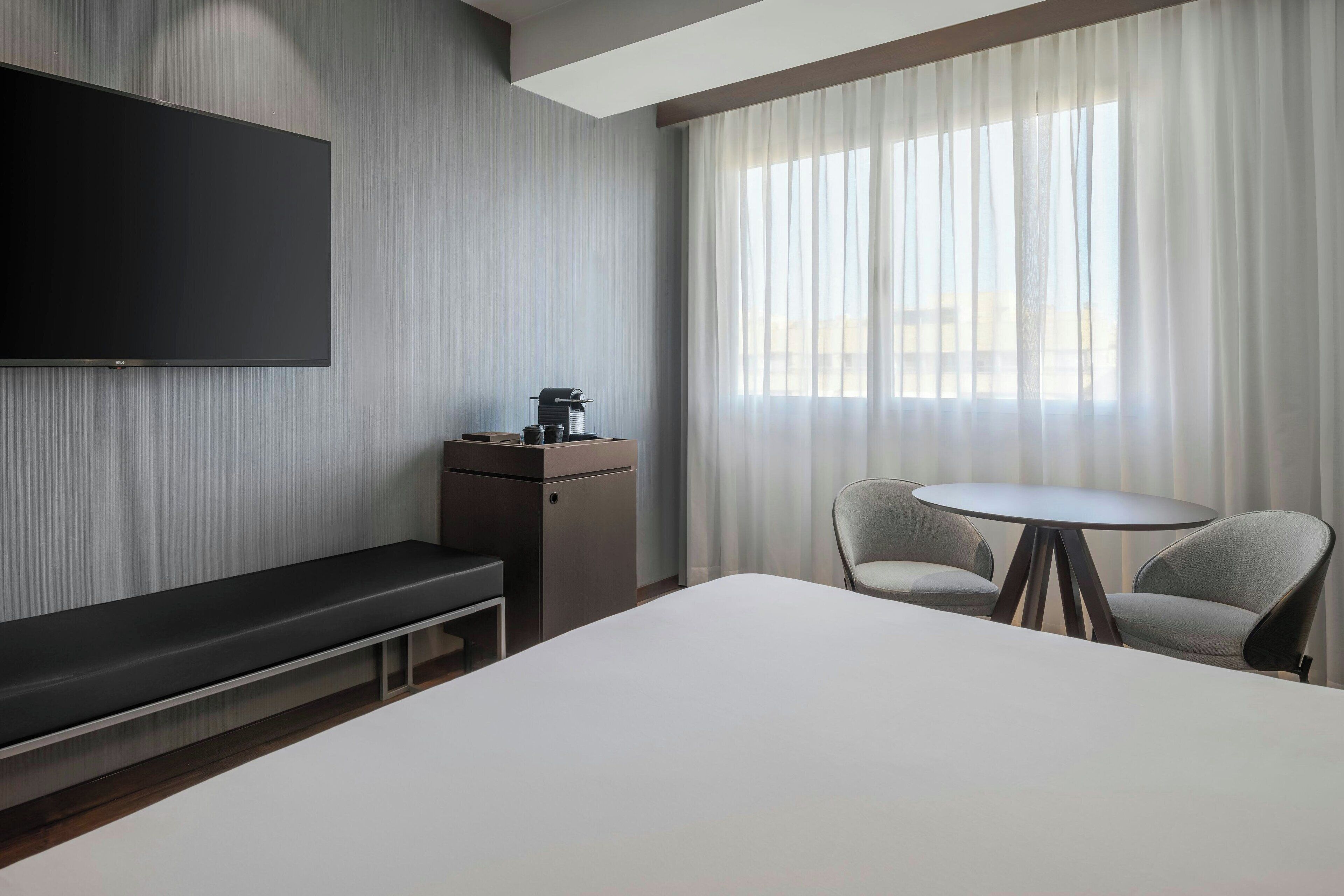 A hotel room with a large bed, a TV mounted on the wall, a small table with chairs, and a window with sheer curtains.