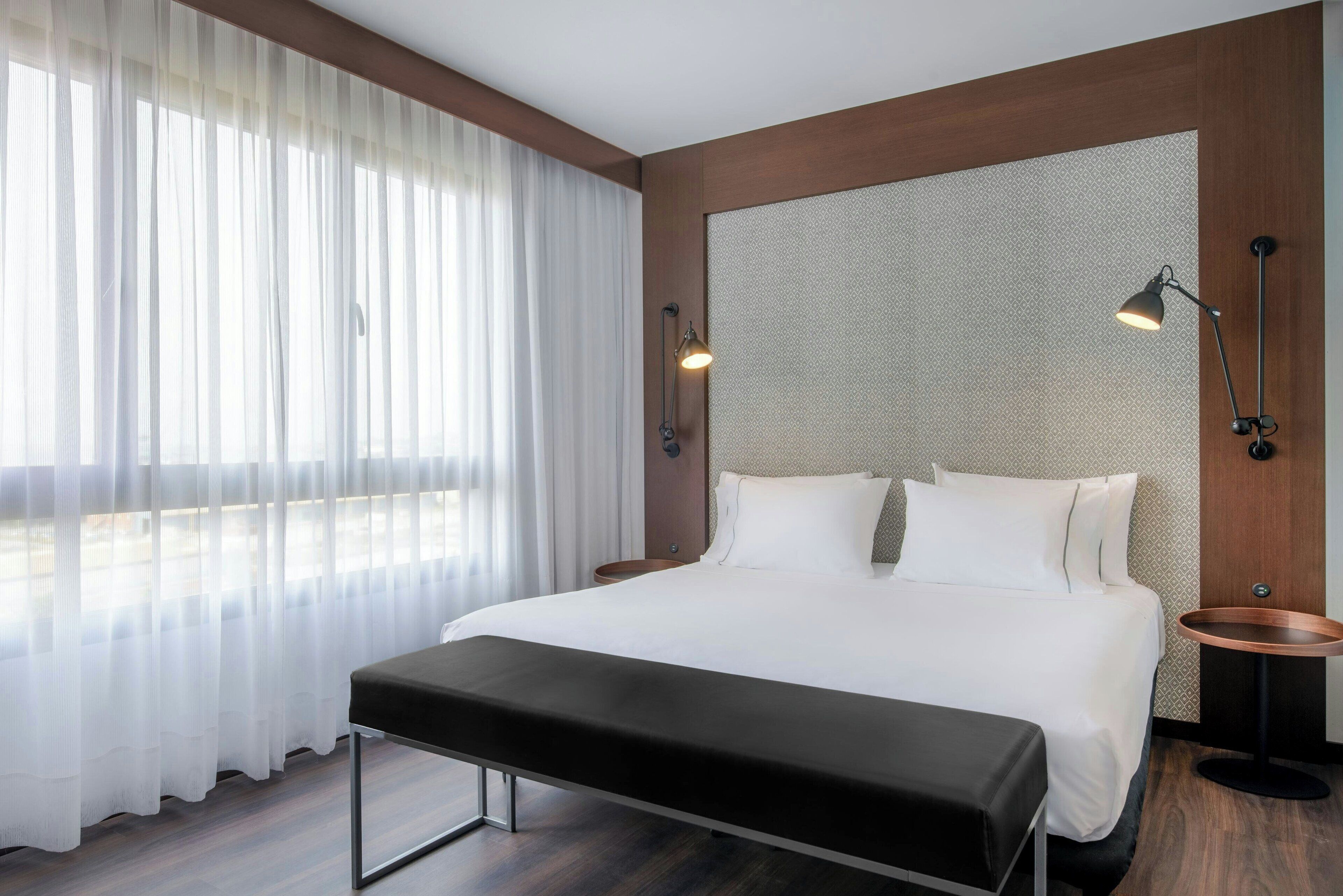 A modern hotel room with a large bed, a bench, a bedside table, and a window with sheer curtains.