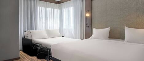 A hotel room with two beds, a large window with sheer curtains, a wall-mounted light, and a wooden floor.