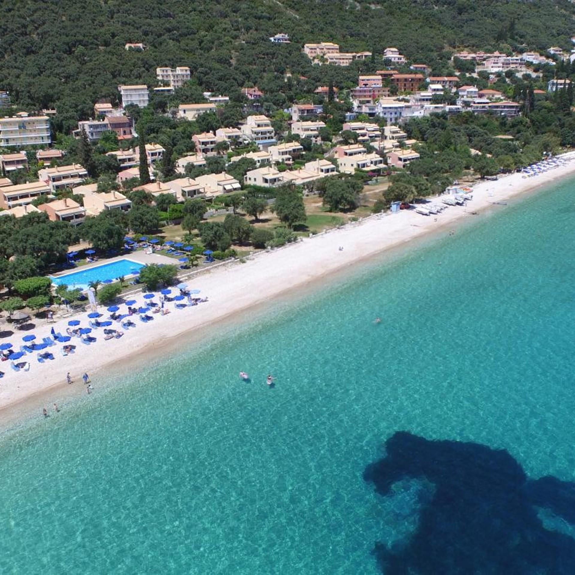 An aerial view of a coastal area with a beach, clear blue water, and a row of buildings with a swimming pool.