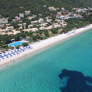 Vue aérienne d’une zone côtière avec une plage, une eau d’un bleu limpide et une rangée de bâtiments dotés d’une piscine.