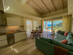 A modern living room with a green sofa, a dining area with a table and chairs, a kitchen with a dishwasher, and a large sliding glass door leading to a balcony with a view of the sea. - La Riviera Barbati (Corfu)