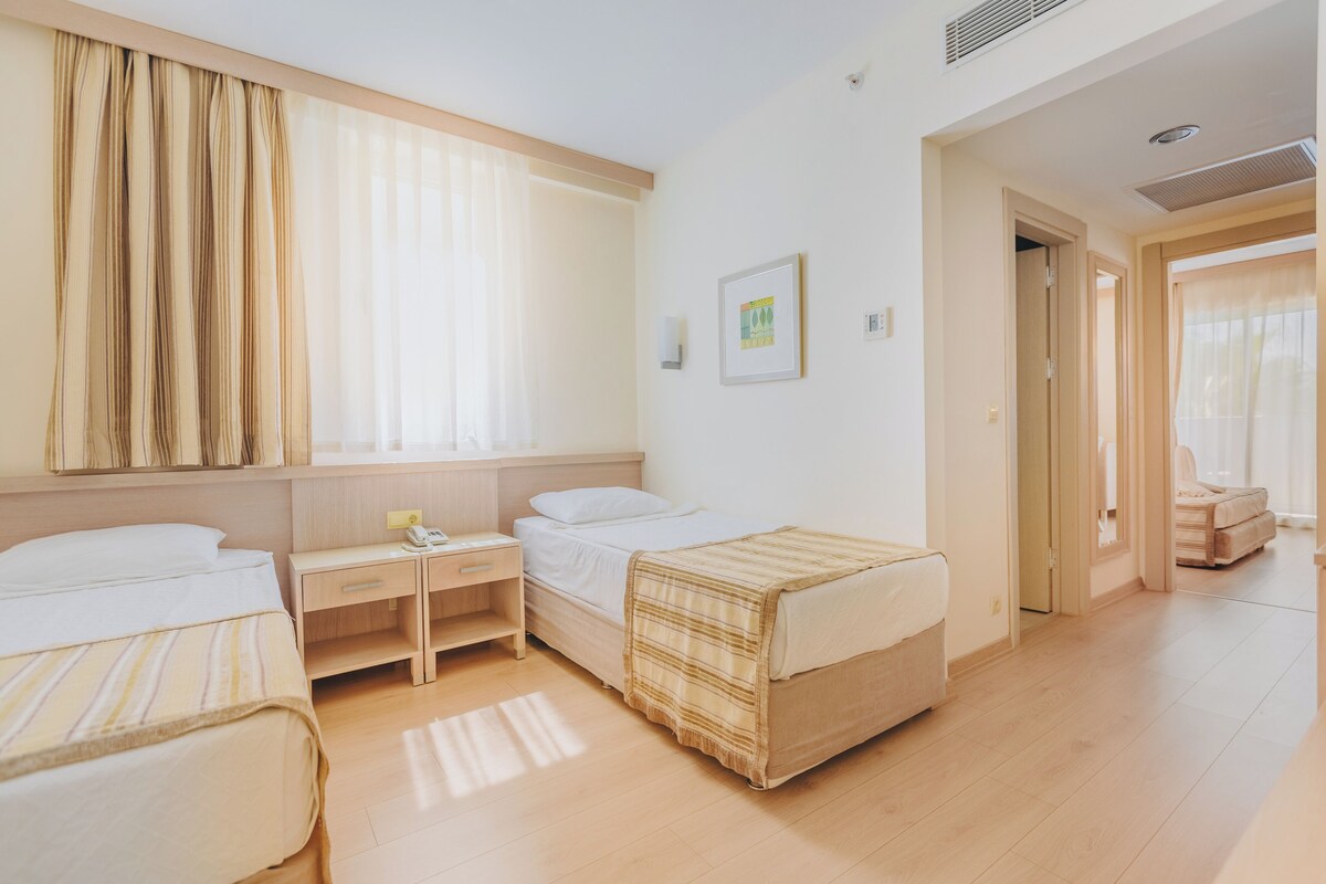 A hotel room with two single beds, a small desk with a chair, a bedside table, and a large window with curtains.